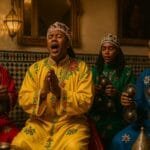 Four Gnawa performers in red, yellow, green, and blue robes playing krakebs in a ceremonial setting.